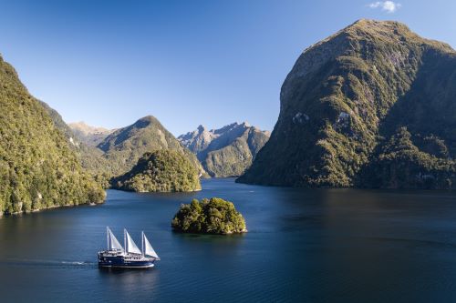 A sailing boat with three sails cruising through Doubtful Sound with the wilderness scenery of steep mountains unfolding in the background