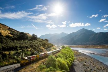 The TranzAlpine train making its way through picturesque New Zealand landscape including mountains, green bush and a small river on a sunny day. 