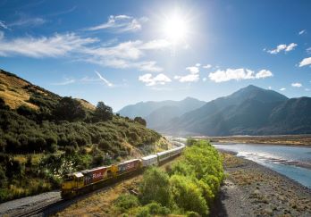 The TranzAlpine train making its way through picturesque New Zealand landscape including mountains, green bush and a small river on a sunny day. 
