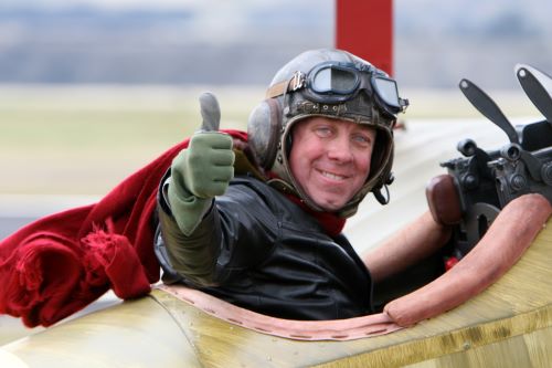 A pilot with a vintage pilot helmet and red scarf sitting in a open canopy plane smiling and giving a thumbs up 