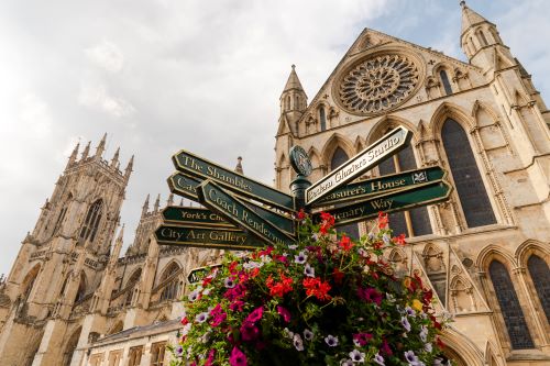 A signpost with blooming flowers hanging of it, indicating directions in all sorts of tourist attractions in York with the cathedral in the background