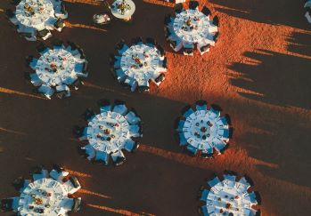 A top down drone image of several round table set for a dinner with white table cloths on the red outback dirt throwing big shadows from the setting sun