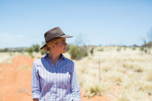 A woman wearing a shirt with stripes and a hat is standing in the outback and looking at the view in the distance