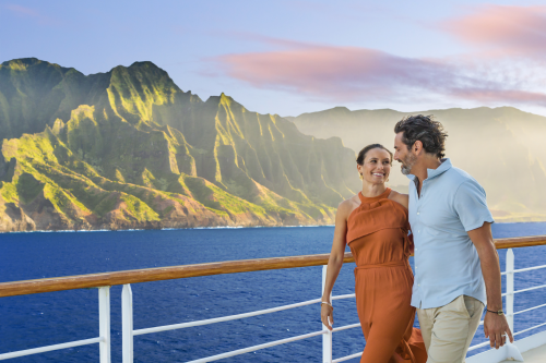 A couple walking aboard the NCL vessel smiling with steep and green Hawaiian mountains behind them.