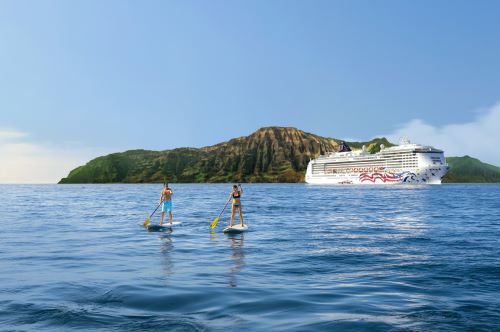 A couple on stand-up paddle boards in the middle of the sea with the NCL vessel and a Hawaiian island in the background. 