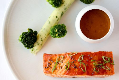 Close up a salmon filet, broccoli and a small bowl with sauce all set up nicely on a plate ready to eat 