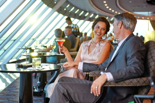 An elegantly dressed couple sitting in a bar drinking a cocktail and smiling with a big panoramic window in front of them to enjoy ocean views from the vessel. 