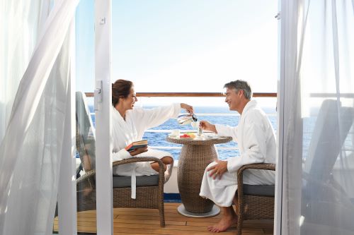 A couple wearing bath robes and sitting on their stateroom's balcony enjoying a breakfast with the wide ocean in the background