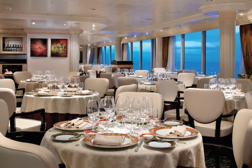 The inside of Oceania's onboard restaurant Toscana with some round tables set for dinner and big windows looking out to the ocean