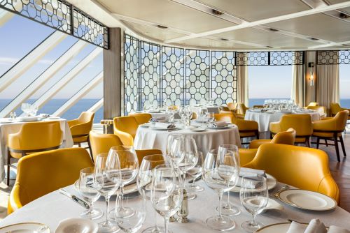 The Toscana restaurant aboard an Oceania vessel with round tables and mustard-yellow leather chairs set up for dining in a contemporary ambience with ocean views