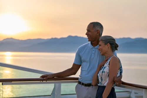 A couple standing at the railing aboard an Oceania vessel and smiling while the sun sets in the background 