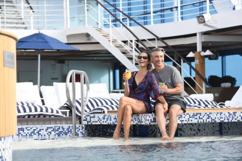 A couple sitting at pool edge with their feet in the water laughing and looking very happy aboard their cruise ship