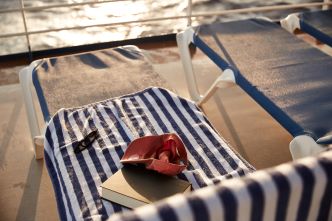 A book, red cap and sun glasses lying on a blue and white striped towel which is covering a sun lunge on board of the cruise ship