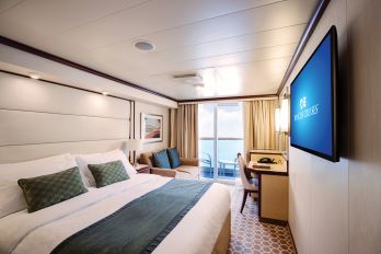 The deluxe balcony stateroom with a big comfortable bed and a TV on the wall, a sofa and study in the back of the room and big windows leading to the balcony 