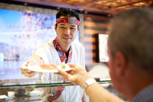 A sushi chef offering a plate of fresh sushi to a passenger