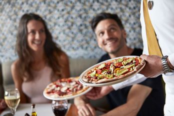 A waiter is serving two plates of delicious pizza to a smiling couple 