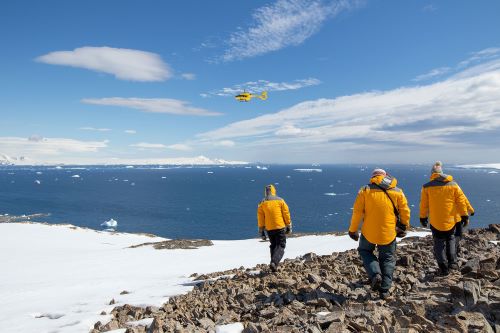 Passengers in the bright yellow parkas walking on icy ground during a heli landing with the Quark heli in the air above them