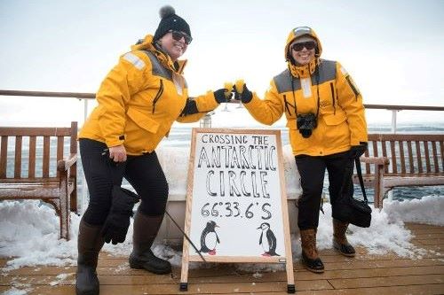 Two travellers in yellow Quark Expedition parkas are celebrating their Antarctic circle crossing by toasting with a drink and smiling in the camera. 