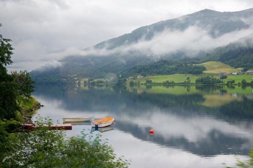 A serene lakeside scene with small boats floating on calm water, surrounded by misty mountains and lush greenery.