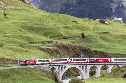 A bright red Glacier Express train crossing a scenic bridge amid lush green hills in Switzerland.