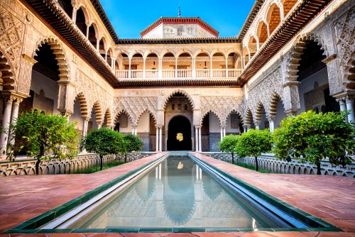 Symmetrical photo of the stunning palace complex Real Alcazar with intricate Moorish architecture, colorful tilework and a long basin. 