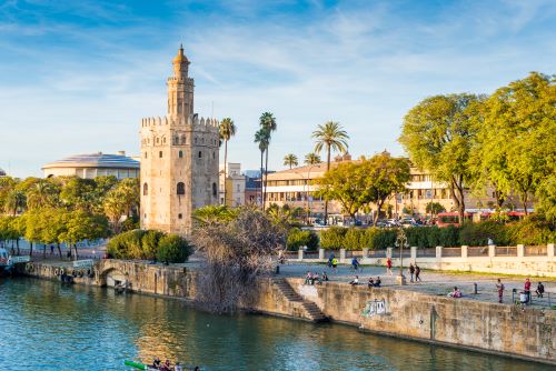 A tall, round, golden-hued stone tower by the river in Seville.