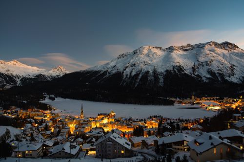 A picturesque snowy town nestled in the Alps with warmly lit buildings surrounded by majestic mountains.