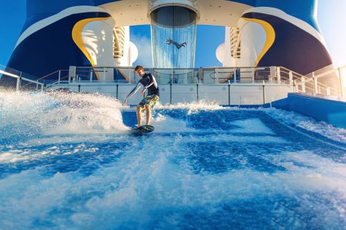 A person surfing on an artificial wave aboard a cruise ship, with a skydiving simulator and someone floating inside it in the background.