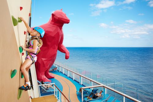 RA girl climbing up a rock wall aboard the vessel with their parent relaxing on a lounge. 