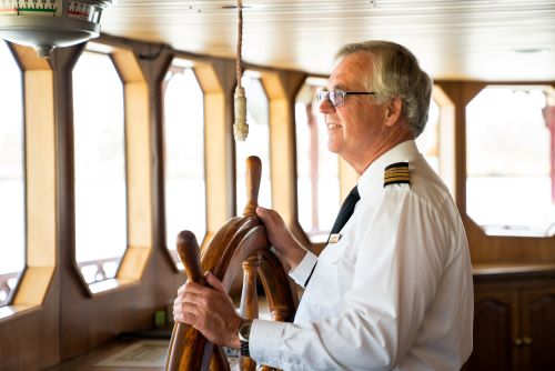 A riverboat captain standing at the helm, steering the boat and gazing forward with a sense of focus and pride.