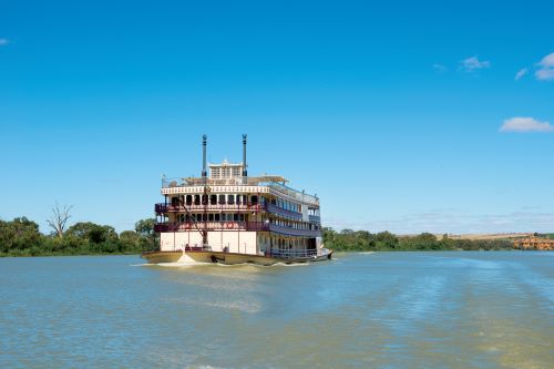 A large, multi-level paddle steamer cruises down a wide, calm river, surrounded by a serene natural landscape under a clear blue sky.