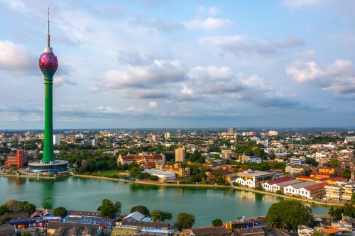 The vibrant cityscape of Colombo featuring a tall green tower with a lotus-shaped top, surrounded by buildings and a reflective lake.