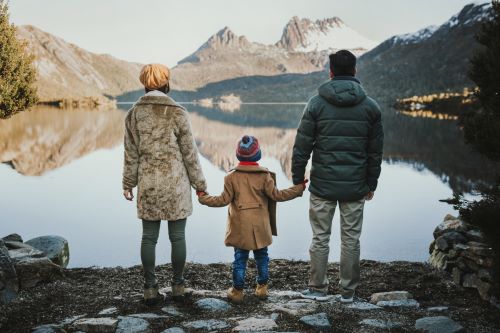 A family of 3 standing with their backs to the camera by the lake at Cradle Mountain with the child in the middle holding the parents' hands