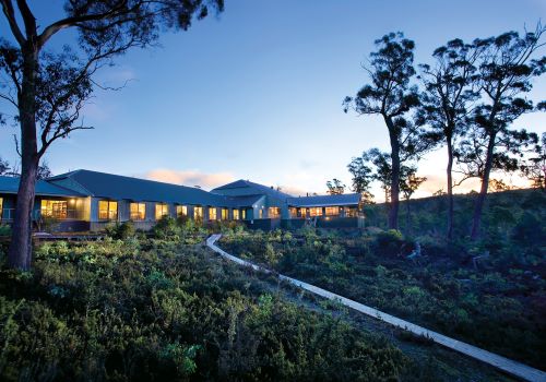 A boardwalk leading to Cradle Mountain Hotel which is lit up towards the sky in twilight