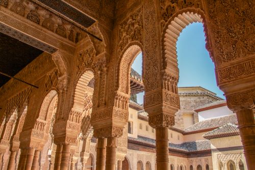 Parts of the intricate architecture of Alhambra Palace captured from beneath a grand ceiling held by massive pillars. 