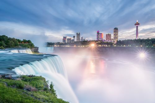 The Niagara Falls cascading down creating lots of mist with the Ontario skyline lit up in the background. 