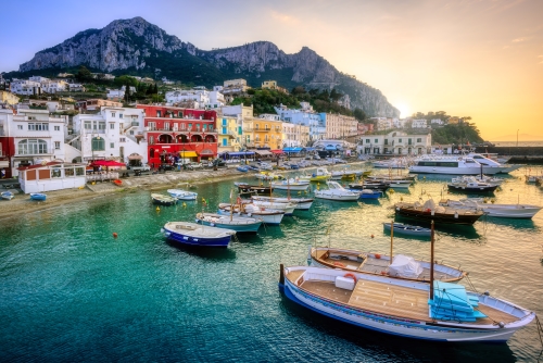 A picturesque harbor with colourful boats on turquoise waters, surrounded by pastel-hued buildings and dramatic cliffs.