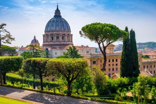 St. Peter’s Basilica with its grand dome rising above lush green gardens, tall cypress trees and elegant historic buildings under a bright blue sky.