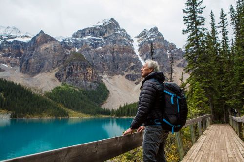 A man with a backpack is standing on a boardwalk looking out to a crystal blue lake and surrounded by steep snow-capped mountains