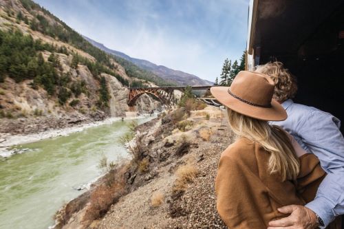 A couple aboard the Rocky Mountaineer train is looking outside from the train's viewing platform and taking in the view of the mountains and river