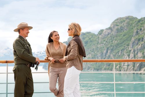 Two female passengers are talking to a guide in uniform on deck of the Holland America Line vessel with some mountains in the background