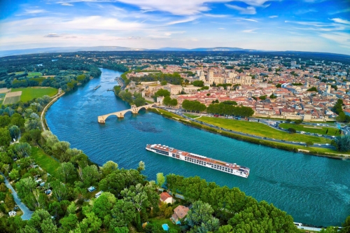 An aerial view of the Viking vessel going down a winding river passing a European town