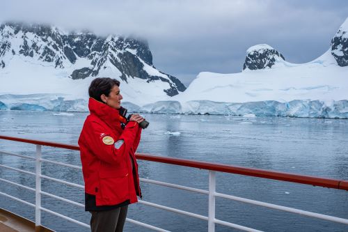 A cruise guest dressed in a red snow jacket holding binoculars looking out from a ship deck to the Antarctic landscape 