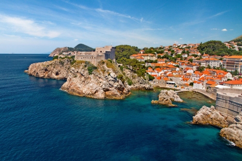 A coastal town with red-roofed buildings nestled around a rocky shoreline, featuring a large fortress on a cliff overlooking the clear blue sea.