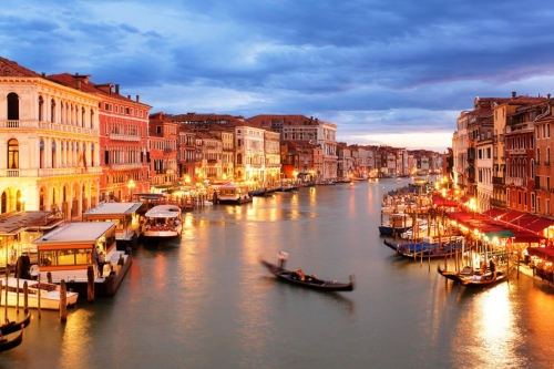 A vibrant canal in a city at twilight, lined with illuminated buildings and bustling with gondolas and boats.