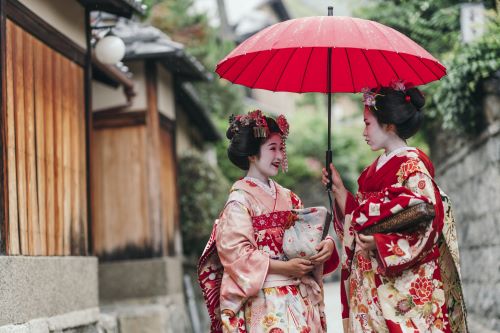 Two Geishas in traditional robes standing under a parasol chatting to each other 