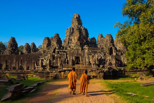 Two monks in bright orange robes walking towards the ancient stone faces of Bayon temple. 