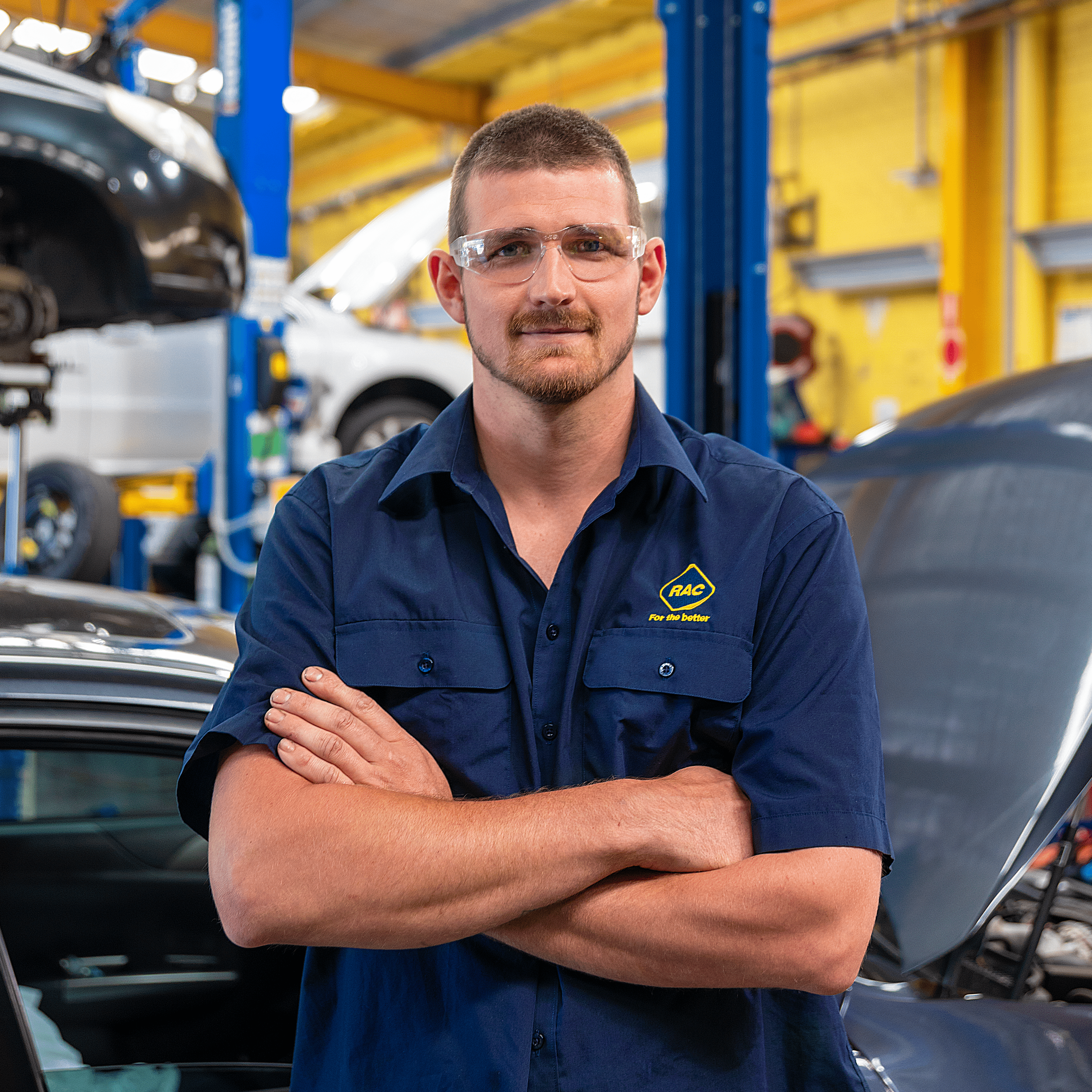 An RAC auto mechanic is standing in front of a car with his arms crossed, smiling