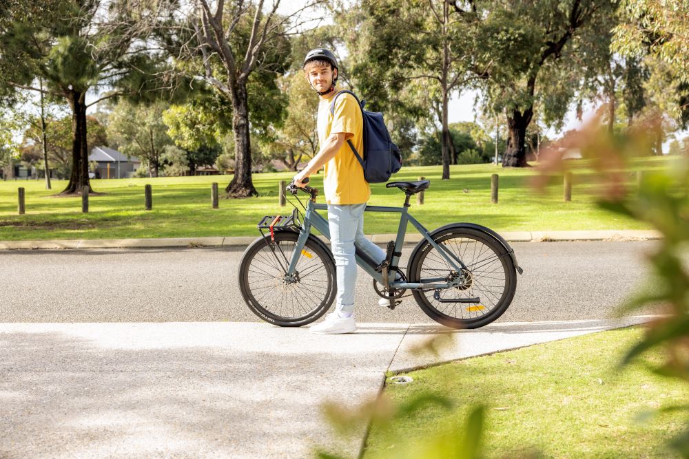 A young man rides an electric bike on the footpath of a leafy street on a sunny day