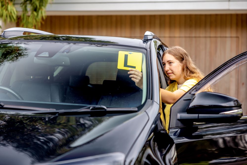 A teen places a yellow L plate sticker in the front window of their family car